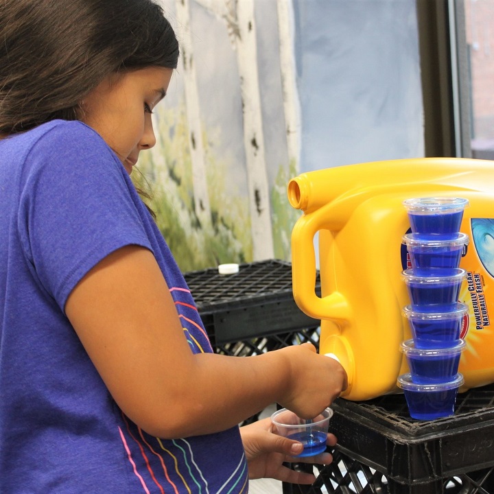 girl filling portion cup with laundry detergent, 6 more stacked nearby