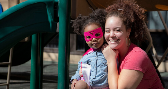 Close-up: woman smiling. She's crouched down next to her daughter, who's wearing a pink costume mask