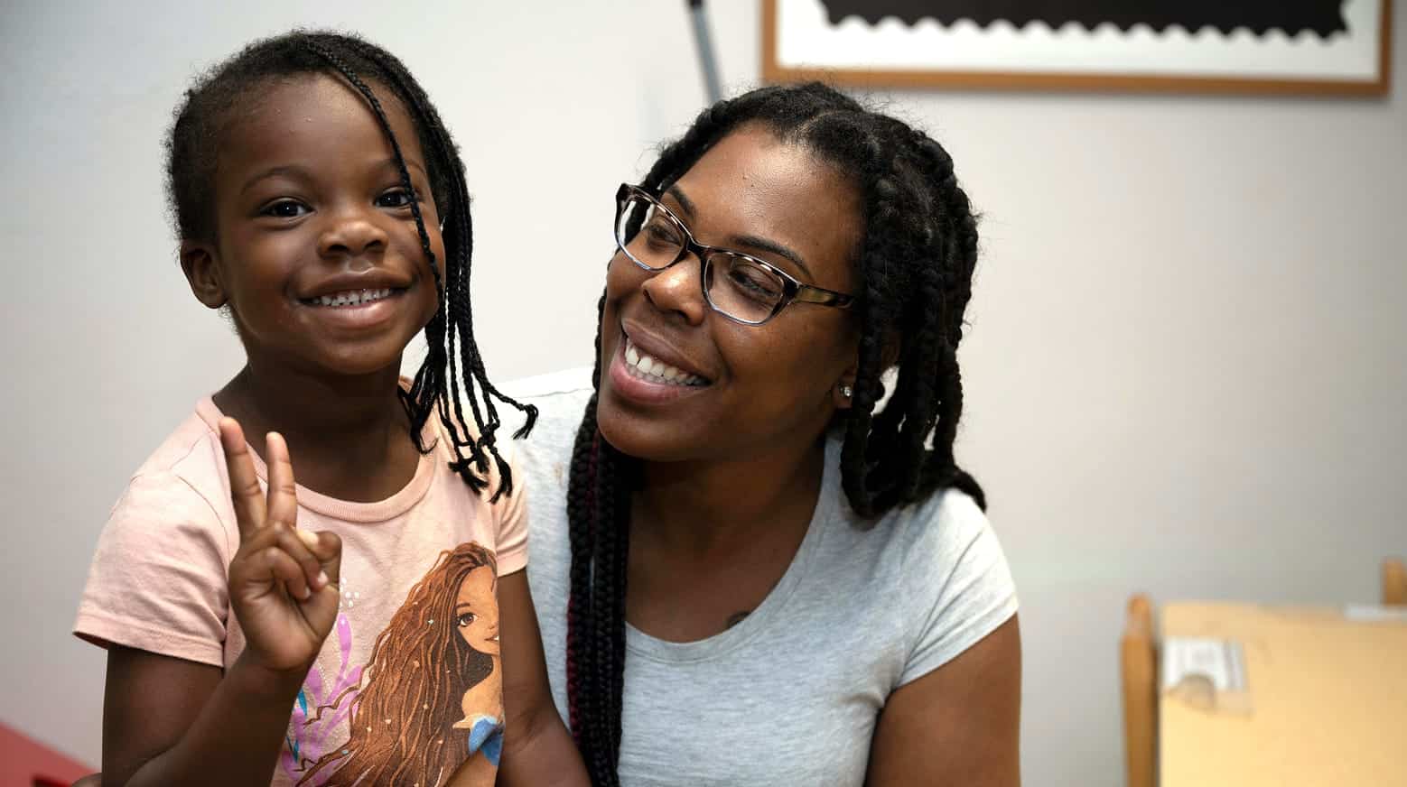 Smiling woman with glasses looks at a young girl who is smiling and holding up a peace sign in a classroom setting.