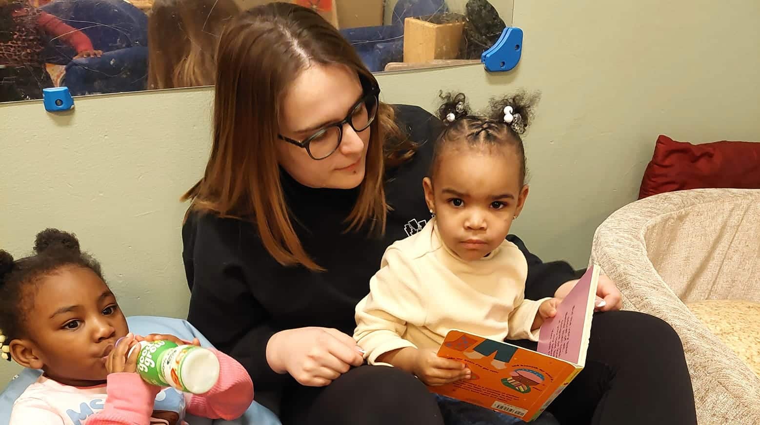 A woman reads to a young girl holding a book, while another girl drinks from a bottle beside them.