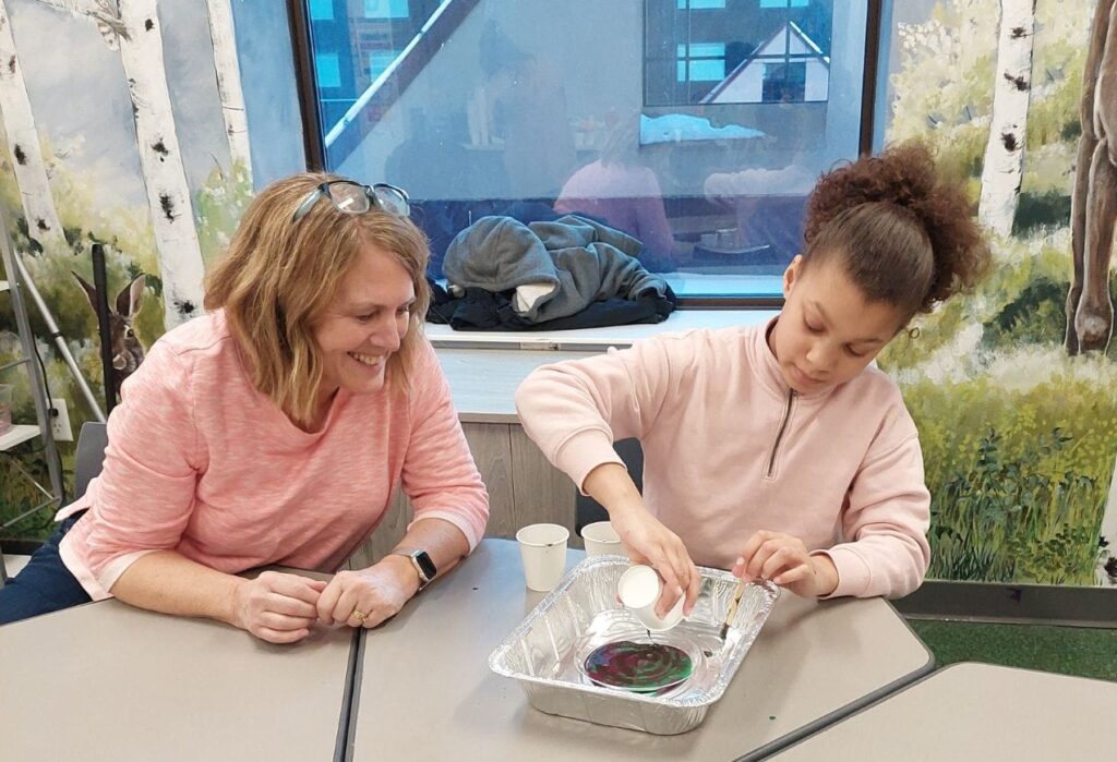 A woman smiles as a young girl pours liquid into a pan during a science activity at a table indoors.