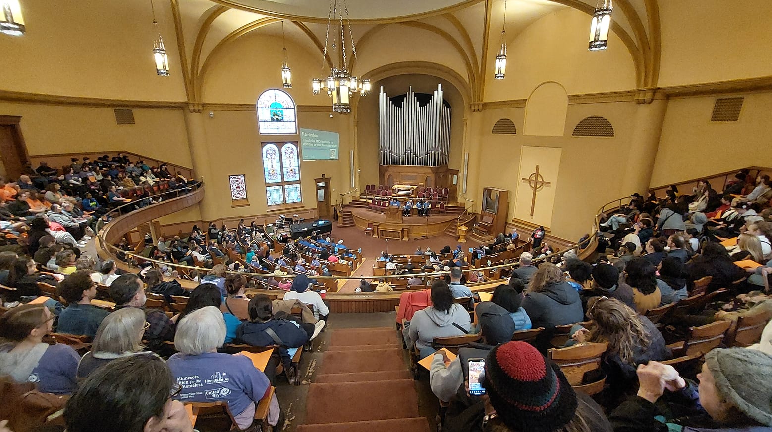 A large audience sits in a church auditorium, listening to speakers on stage near an organ and stained glass windows.