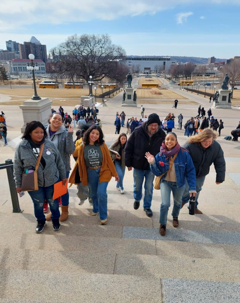 A group of people smiling and climbing outdoor stone steps with a cityscape and crowd in the background.