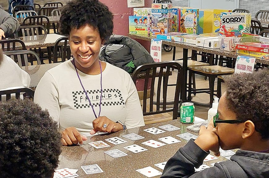 A woman and two children play a card game at a table, with board games and drinks visible in the background.