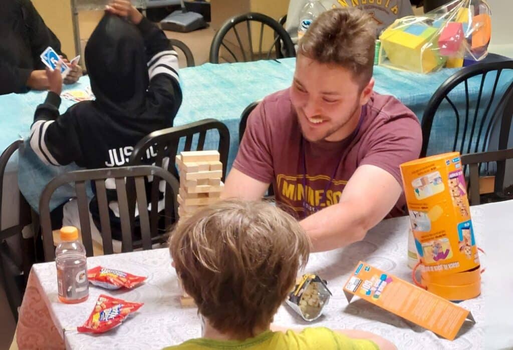 Three people play games at a table with snacks; one smiles and plays Jenga while others play UNO in the background.