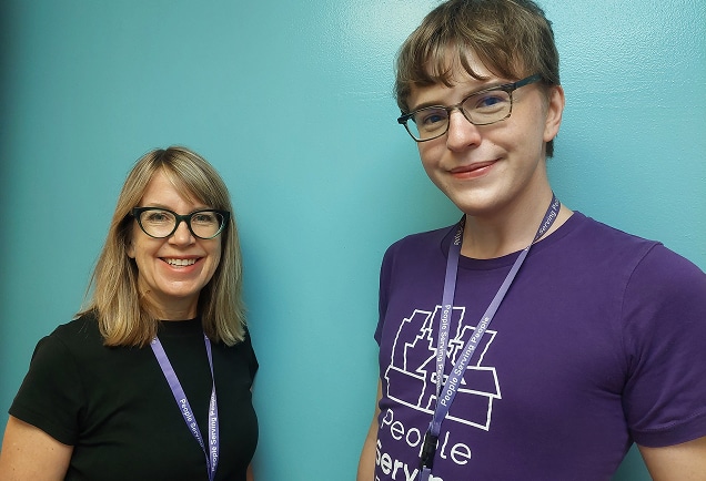 Two people wearing glasses and lanyards stand smiling in front of a bright blue wall.