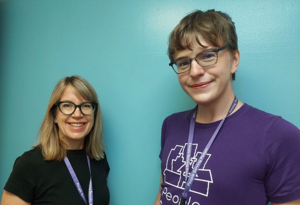 Two people wearing glasses and lanyards stand smiling in front of a blue wall.