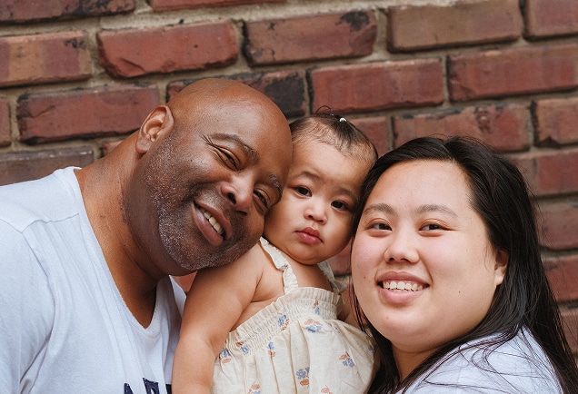 A man, woman, and toddler smile together in front of a brick wall.
