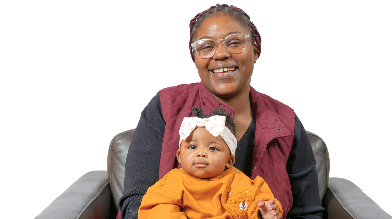 Smiling woman with glasses holds a baby wearing an orange outfit and white bow, both sitting on a chair against a white background.