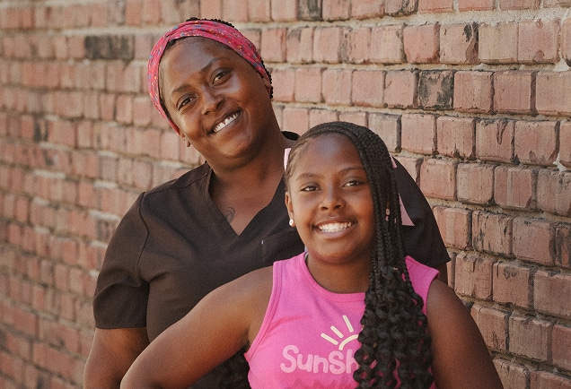 Smiling woman and girl standing close together in front of a brick wall, both looking at the camera.