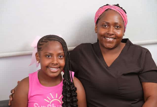A smiling woman sits next to a smiling girl with braids and a pink top.