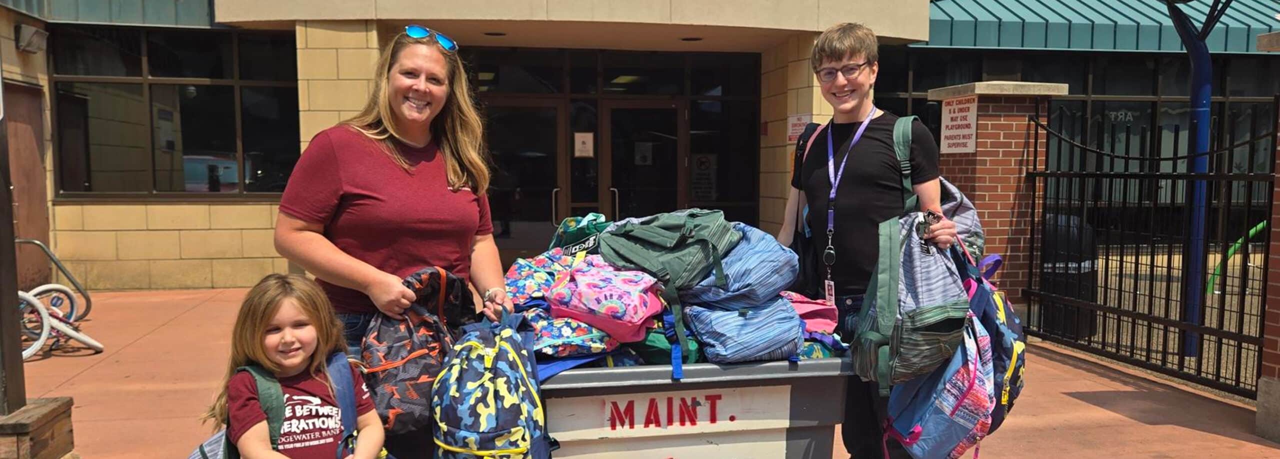 Three people stand outside a building, smiling and holding backpacks near a cart piled high with colorful backpacks.