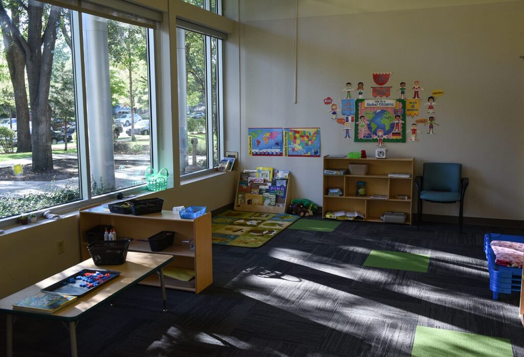 Sunny classroom with large windows, bookshelves, maps, toys, and colorful educational posters on the wall.