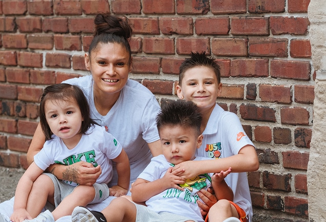 A woman and three young children in white shirts sit smiling together by a brick wall.