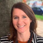 A woman with long brown hair smiles outdoors, wearing a striped jacket and a chunky black necklace.