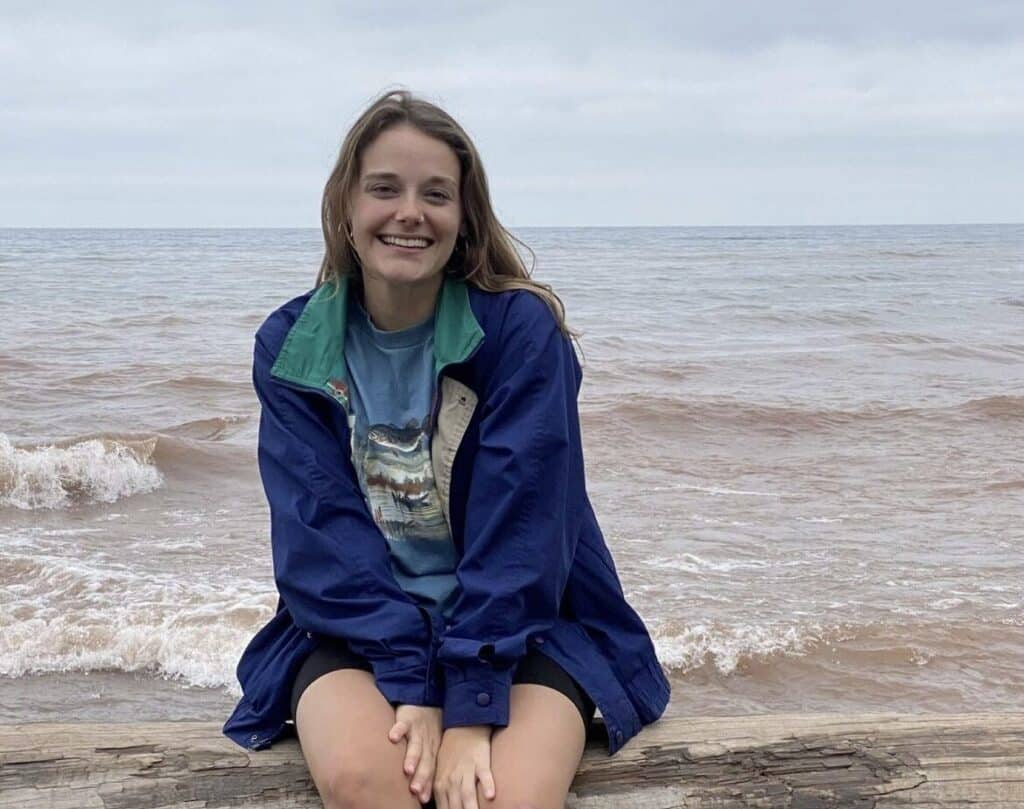A young woman smiles while sitting on a log at the beach, with waves and a cloudy sky in the background.