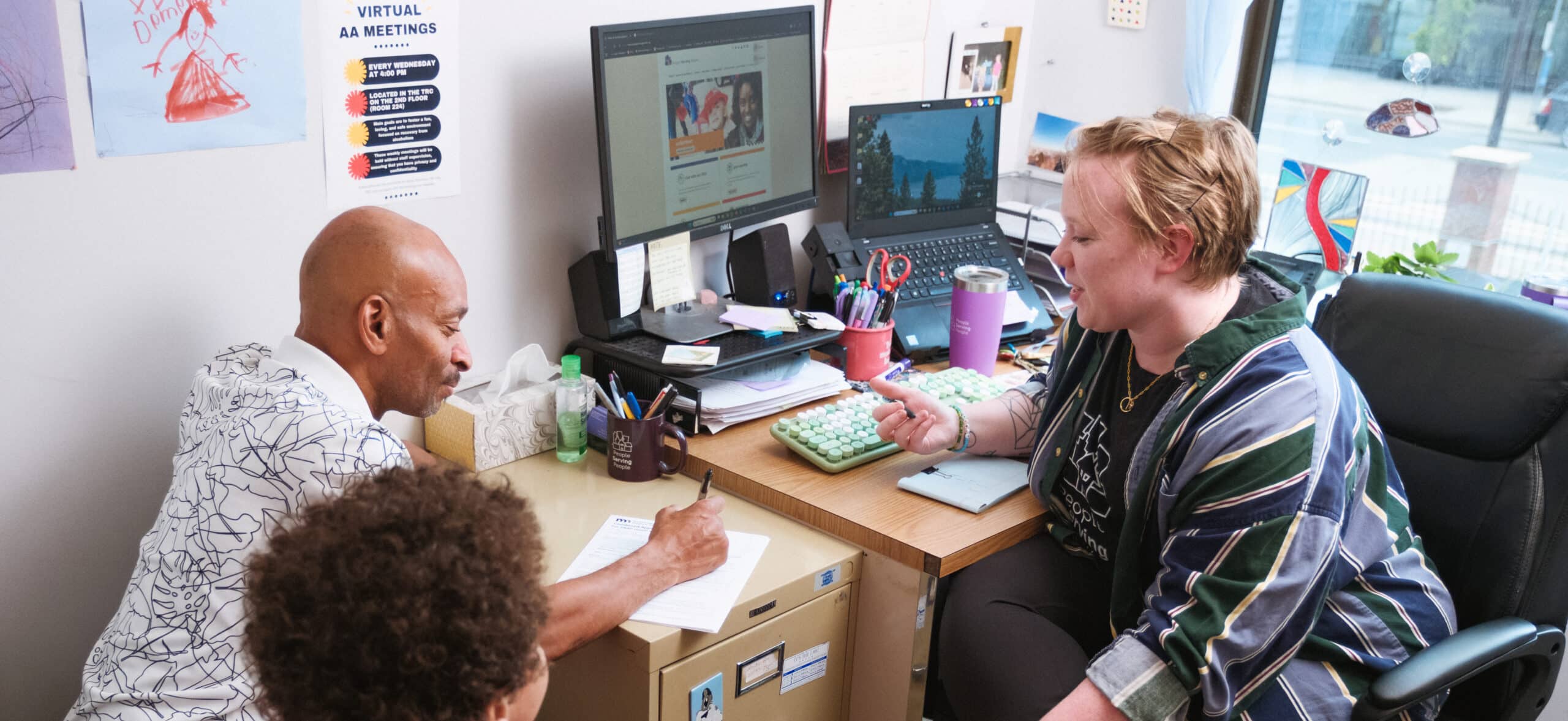 Two adults and a child talk together in a small office with computers, papers, and colorful artwork on the walls.