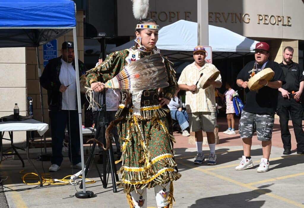 A Native American dancer in regalia performs outdoors as two men play hand drums; a crowd watches nearby.