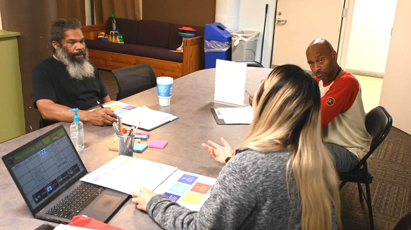 Three people sit around a table with laptops and papers, engaged in a discussion in a casual office setting.