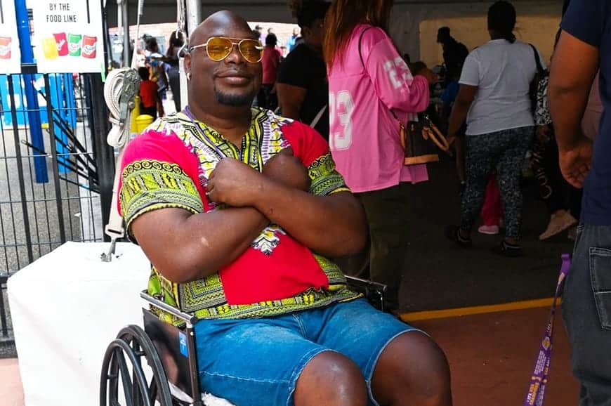 A man in a wheelchair, wearing a colorful shirt and shorts, smiles and crosses his arms at an outdoor event.
