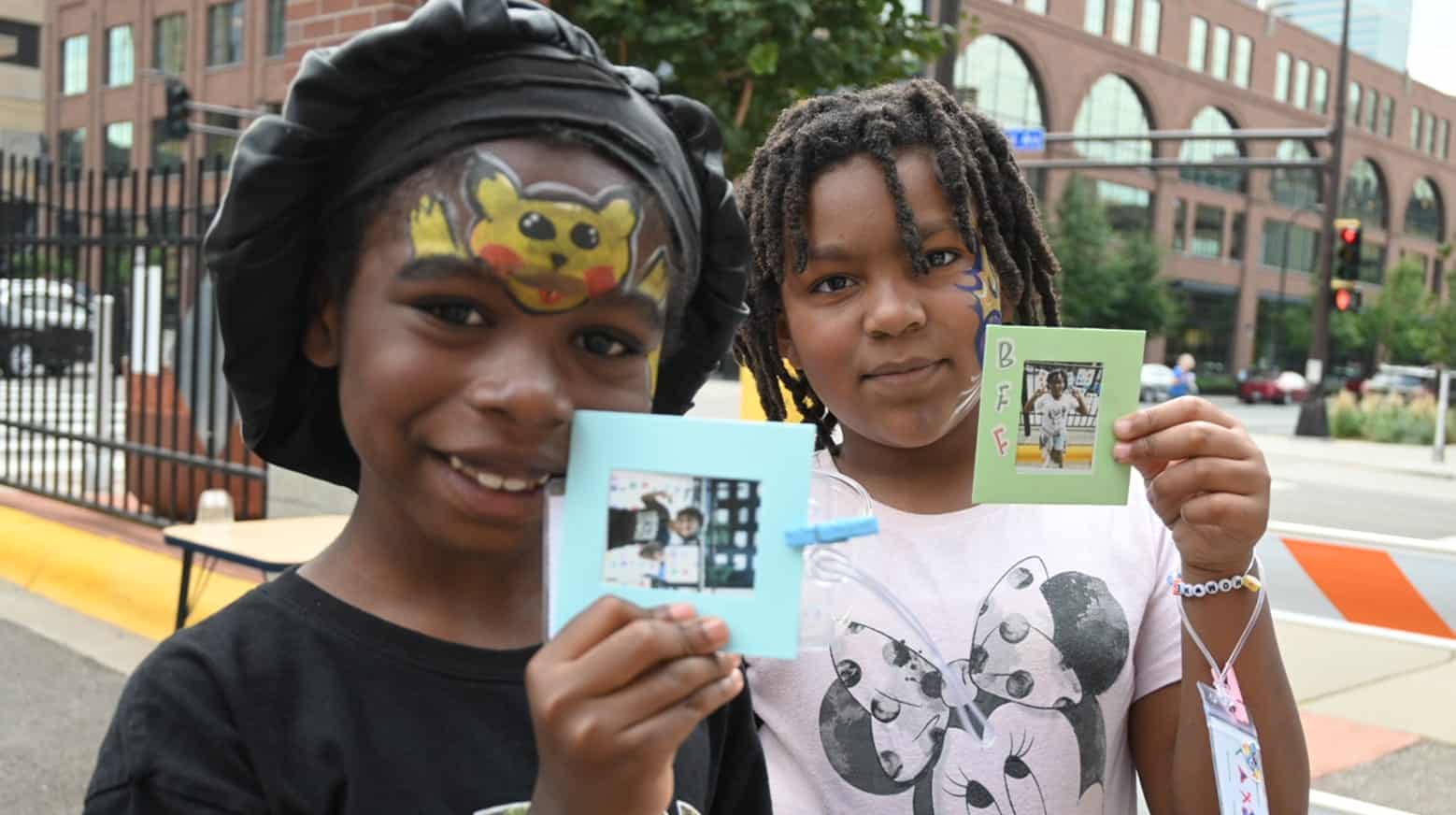Two smiling children with face paint hold up decorated photos outdoors in a city setting.