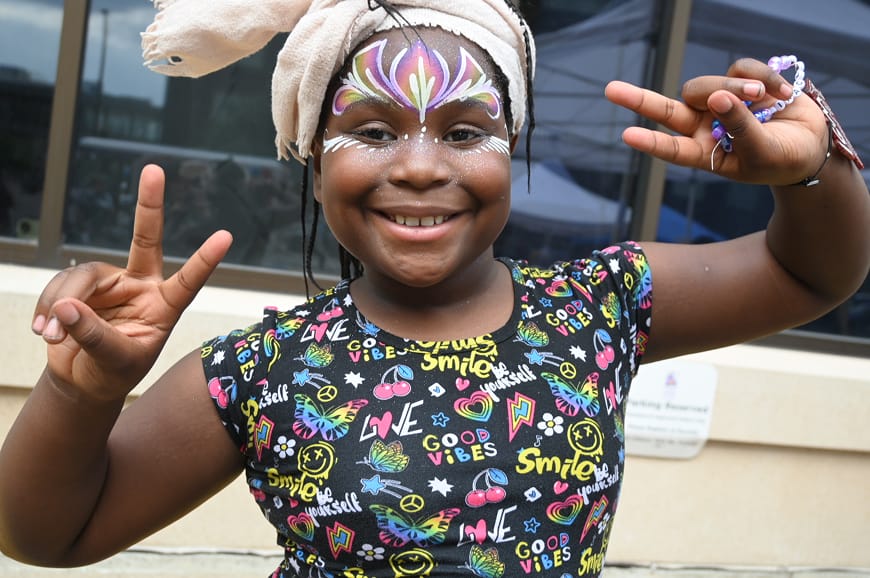 Smiling child with colorful face paint and a headwrap makes peace signs with both hands, wearing a "Good Vibes" shirt.