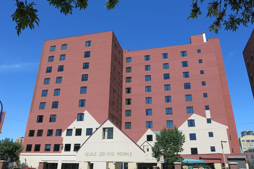 A tall red-brick building with white sections at the base, labeled "People Serving People," under a clear blue sky.
