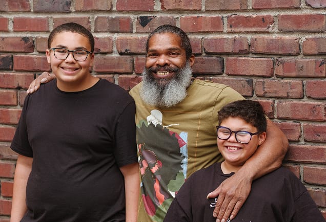 Smiling man with a beard stands by two boys in glasses, all posing happily in front of a brick wall.