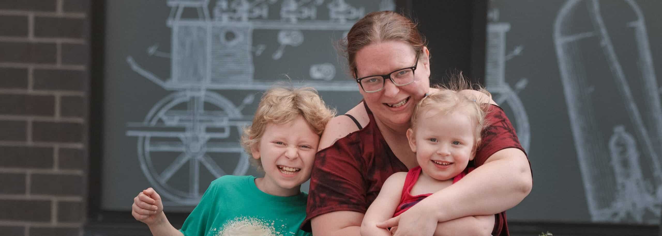 A smiling adult hugs two happy children in front of a chalkboard drawing of a train engine.
