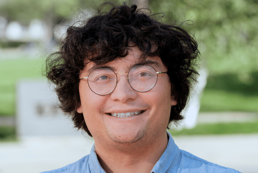 A man with short, wavy dark hair and glasses smiles with greenery in the background.
