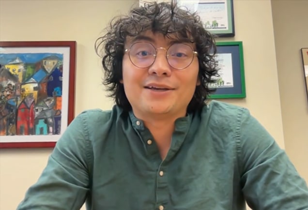Person with curly hair and glasses wearing a green shirt sits indoors, with colorful framed art on the wall behind them.
