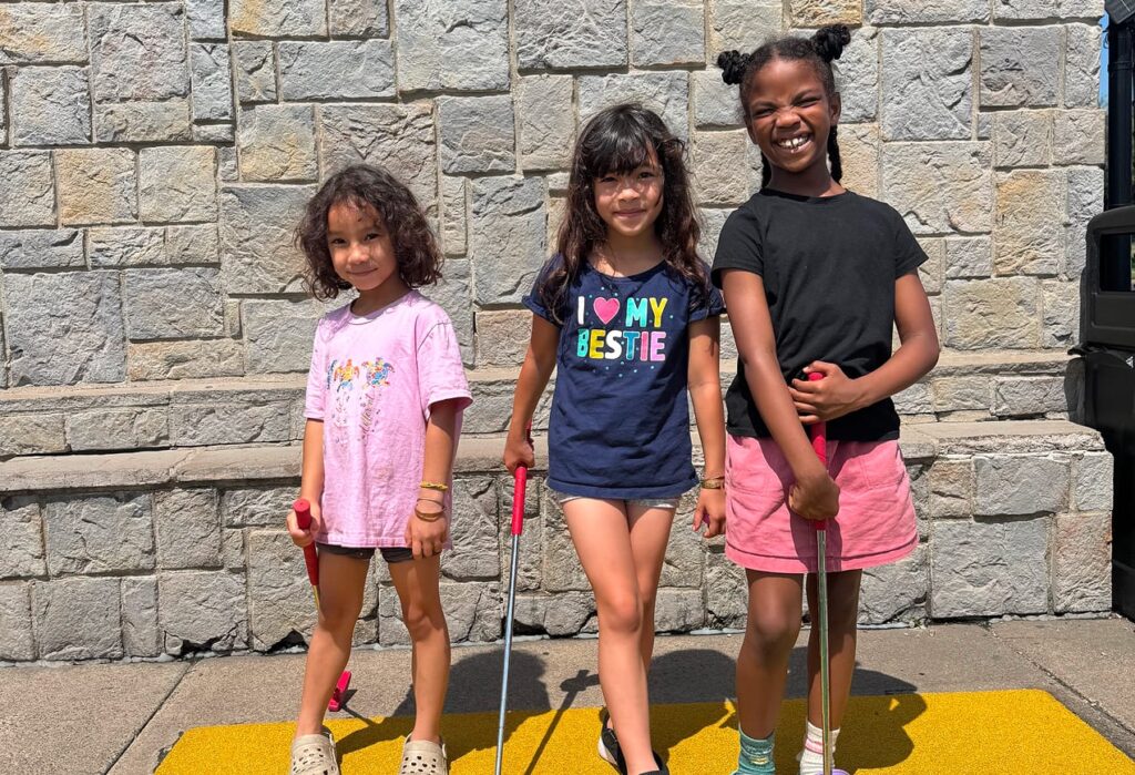 Three smiling young girls holding mini golf clubs stand on a yellow mat in front of a stone wall on a sunny day.