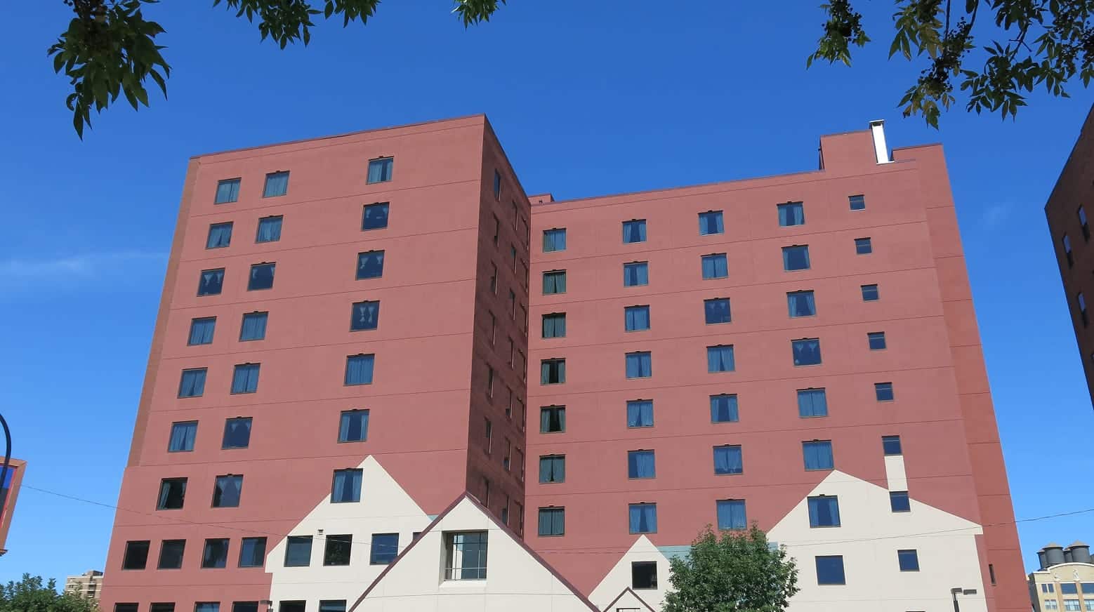 A red-brown apartment building with square windows and white lower sections under a clear blue sky, with tree leaves above.