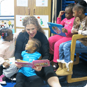 A woman reads a colorful book to four young children, who are smiling and listening attentively in a classroom setting.