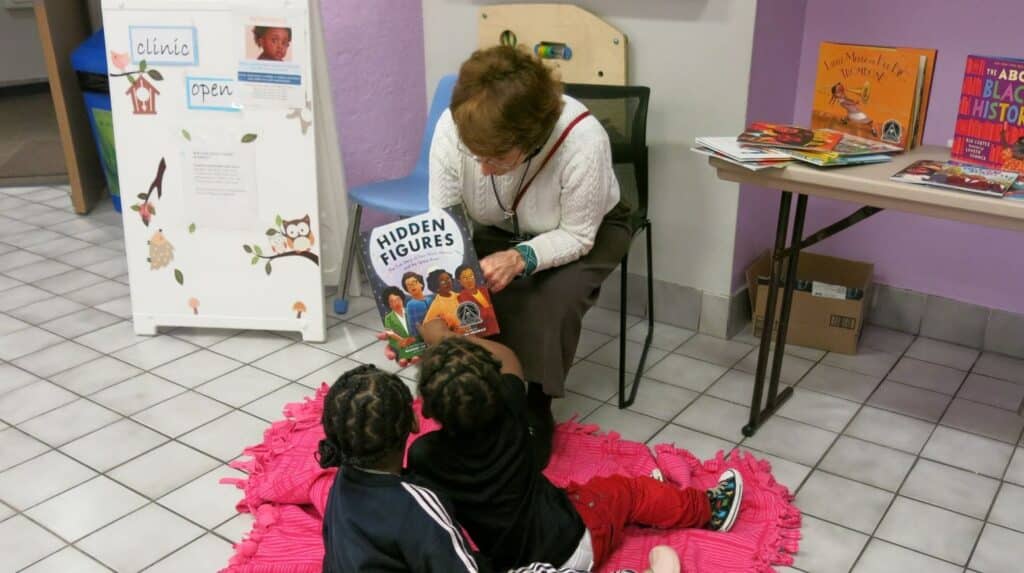 An older woman reads "Hidden Figures" to two young children seated on a pink mat in a cozy reading corner.