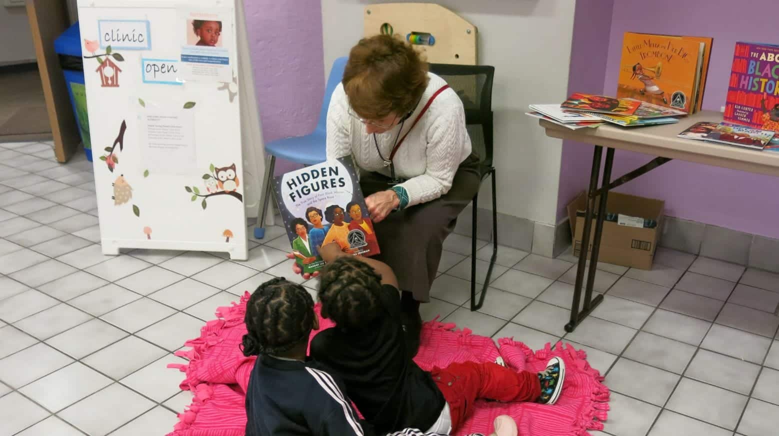 An older woman reads "Hidden Figures" to two young children seated on a pink mat in a cozy reading corner.