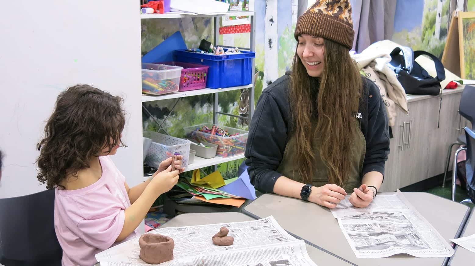 A child and an adult smile while making clay sculptures together at a table covered with newspaper.