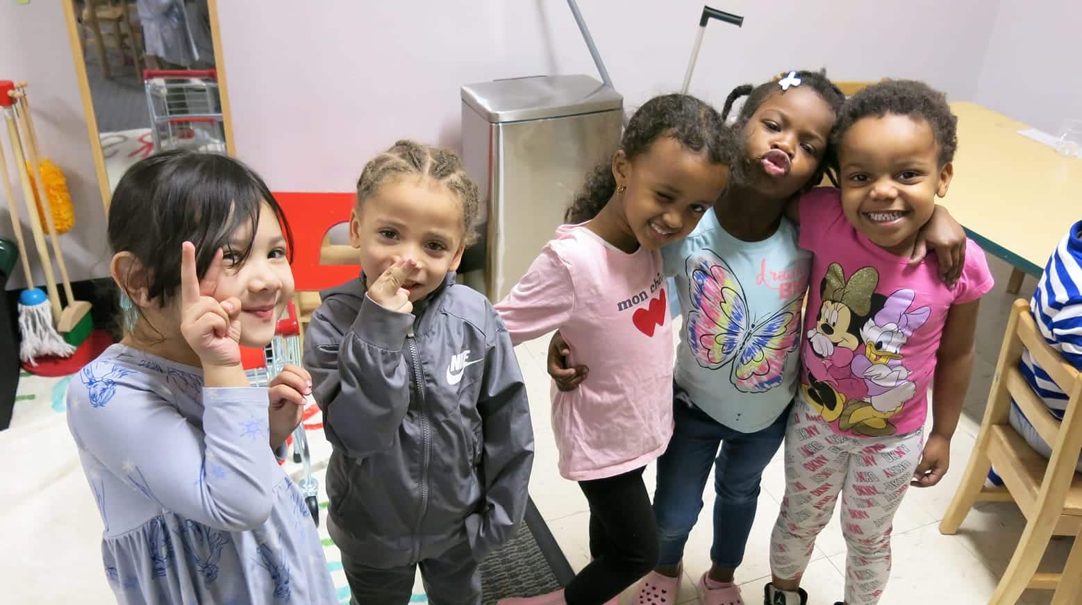 Five young children smile and pose together indoors, some making peace signs, standing close and looking happy.