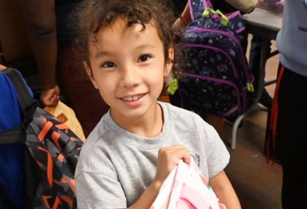 Smiling child holding a pink backpack, surrounded by other children with backpacks indoors.