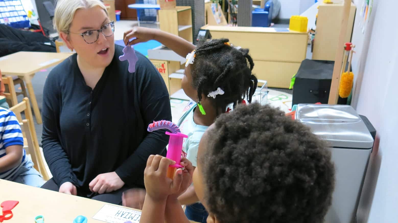 A teacher watches as two young children play with colorful dinosaur toys at a classroom table.