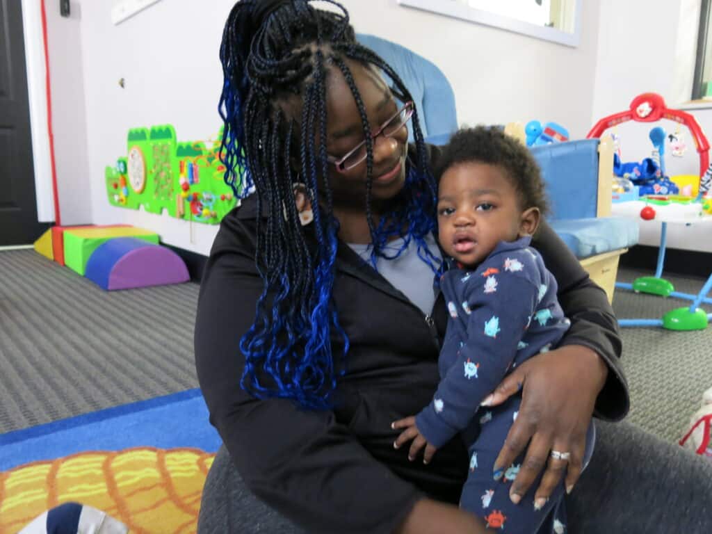 A woman with blue braids holds a baby in pajamas while sitting on the floor in a playroom.
