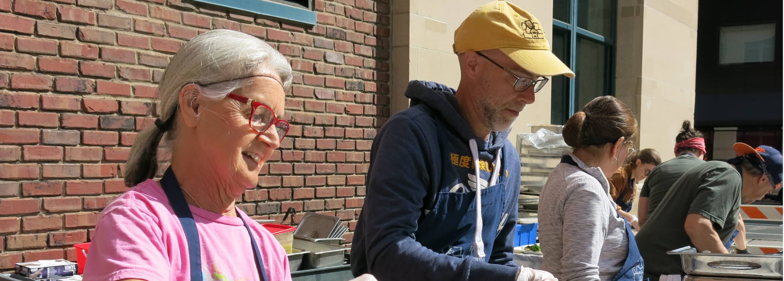Several people stand outside serving food at a table, smiling and working together in the sunlight.