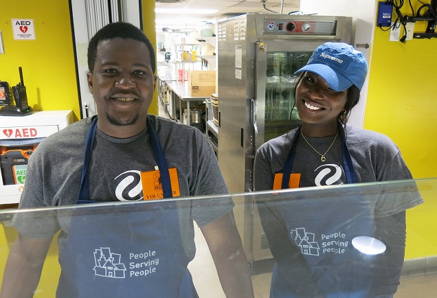 Two smiling volunteers in gray shirts and blue aprons stand behind a counter in a brightly lit kitchen.