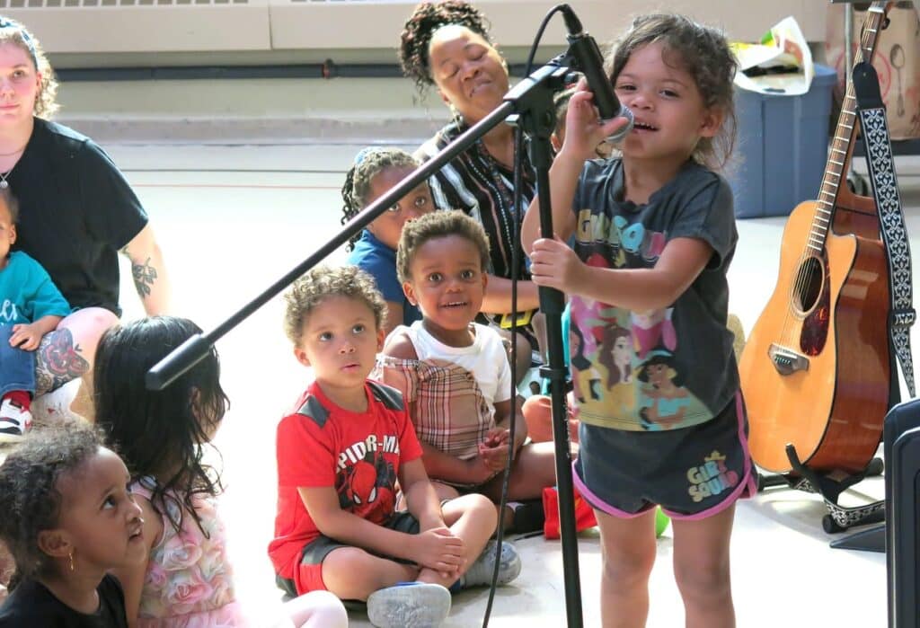 A young girl sings into a microphone while other children sit on the floor watching her in a brightly lit room.