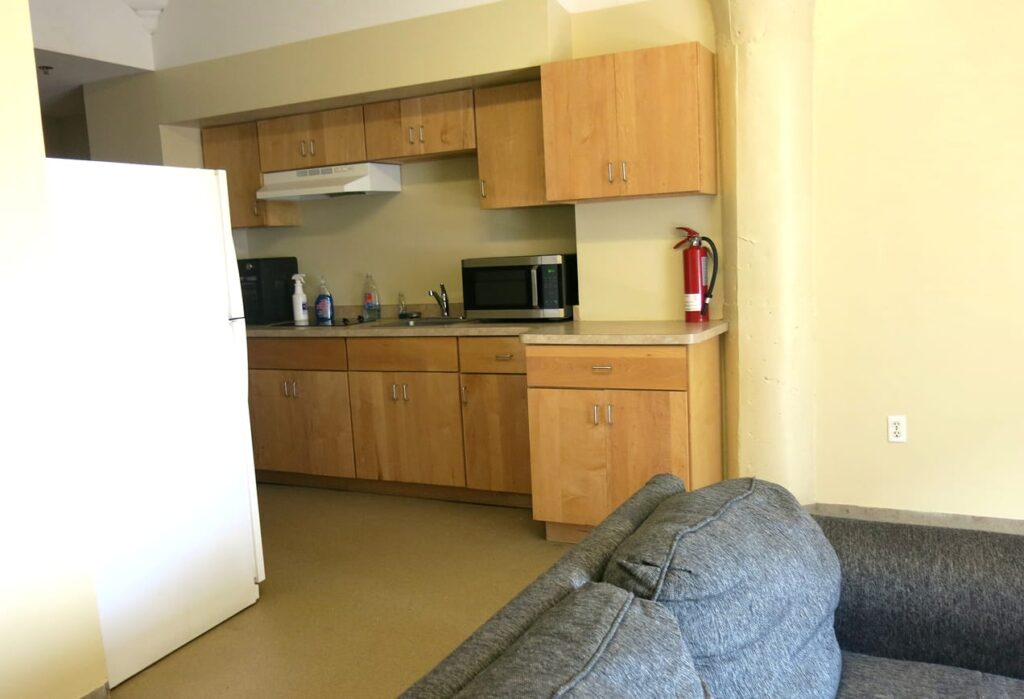 Small kitchen with wooden cabinets, microwave, fridge, sink, cleaning supplies, and a gray couch in the foreground.
