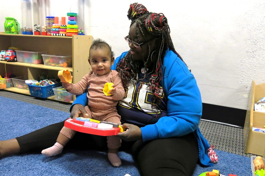A woman and a baby play with toys on a blue carpet in a colorful, child-friendly room.