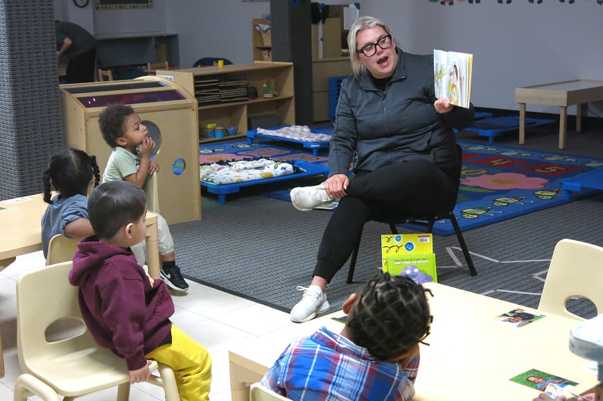 A woman reads a picture book to young children seated at tables in a colorful classroom.