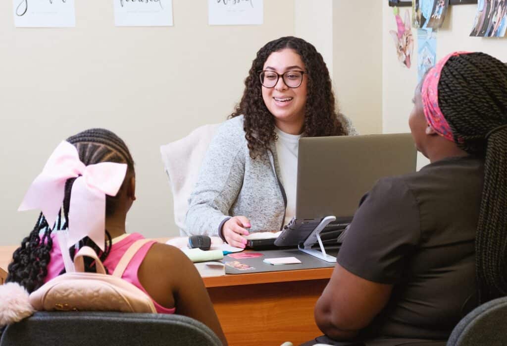A woman at a desk smiles while speaking with a young girl and an adult sitting across from her.