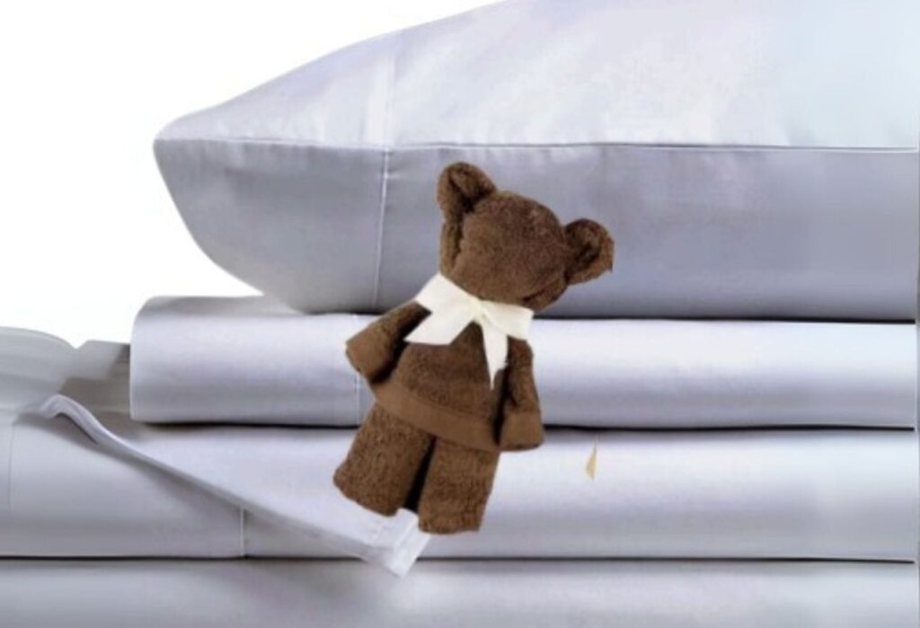 A small brown teddy bear with a white ribbon sits on stacked white pillows and sheets.
