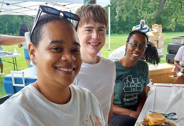 Three people smiling at an outdoor picnic, one holding a plate with a burger, trees and picnic supplies in the background.